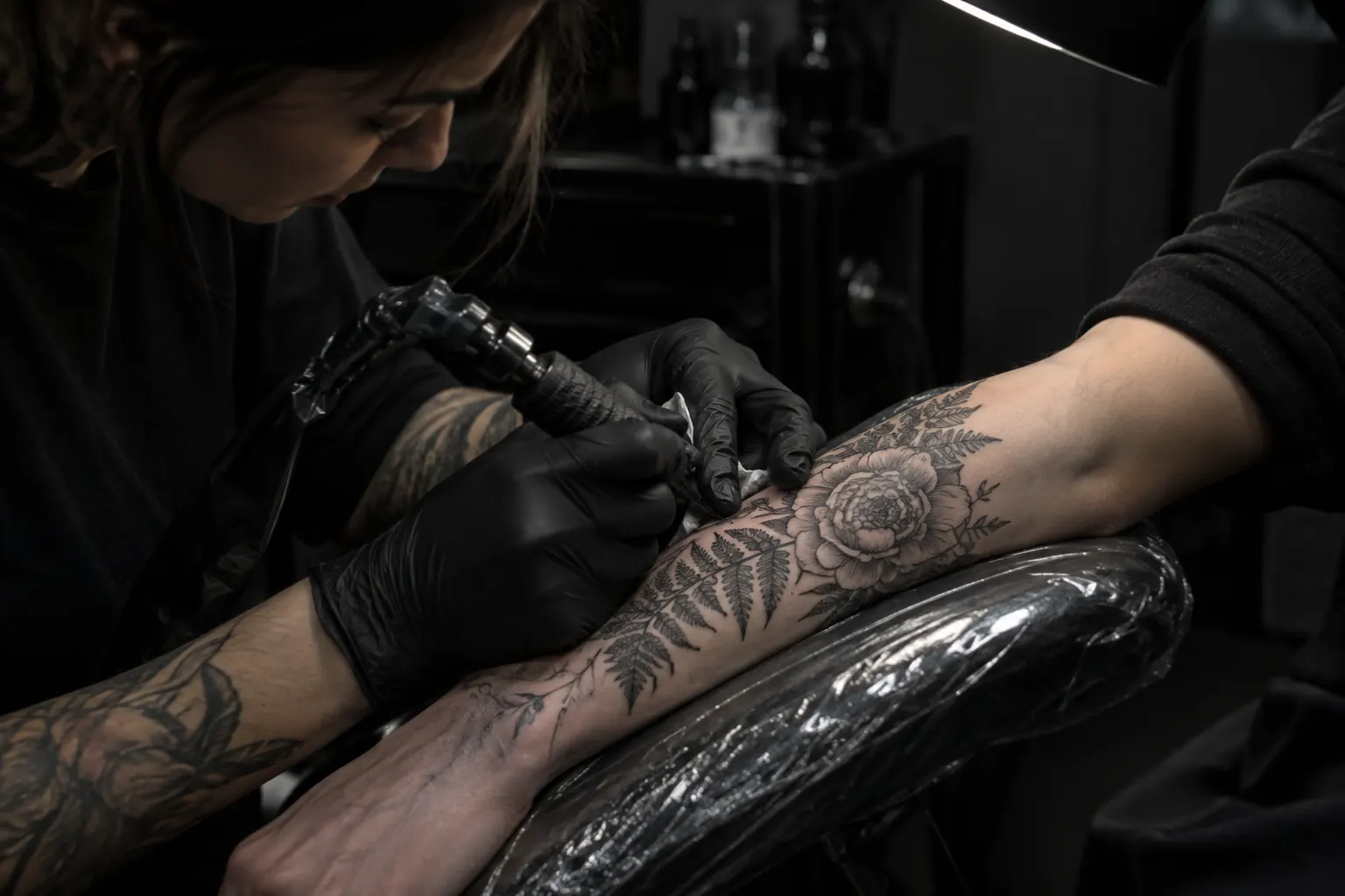 An artist in black gloves tattooing a floral design on a forearm
